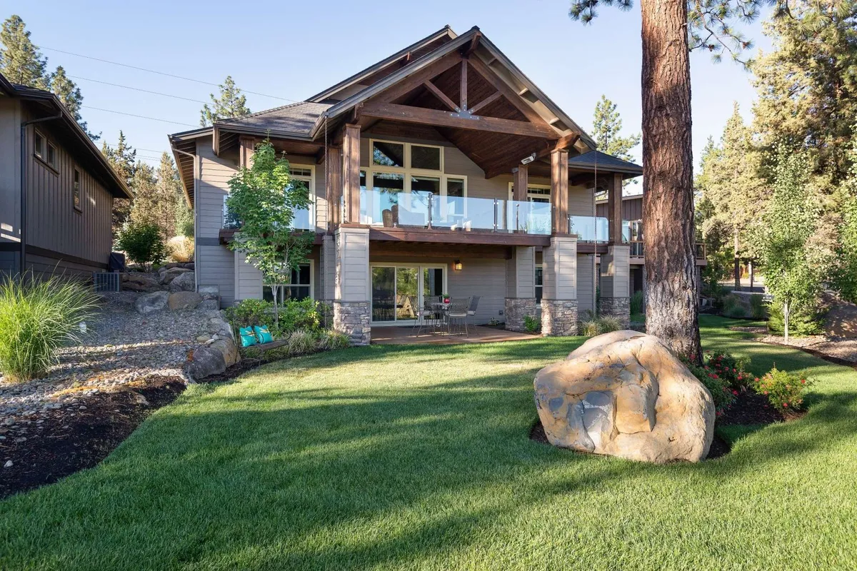 The image features the exterior of WoodRiver House, showcasing a spacious wooden deck overlooking the river, a striking stone facade, manicured gardens, and a large decorative boulder in the foreground. The setting is bathed in soft sunset light, creating a serene and welcoming atmosphere ideal for outdoor gatherings, relaxing with coffee, or stargazing under clear Oregon skies.