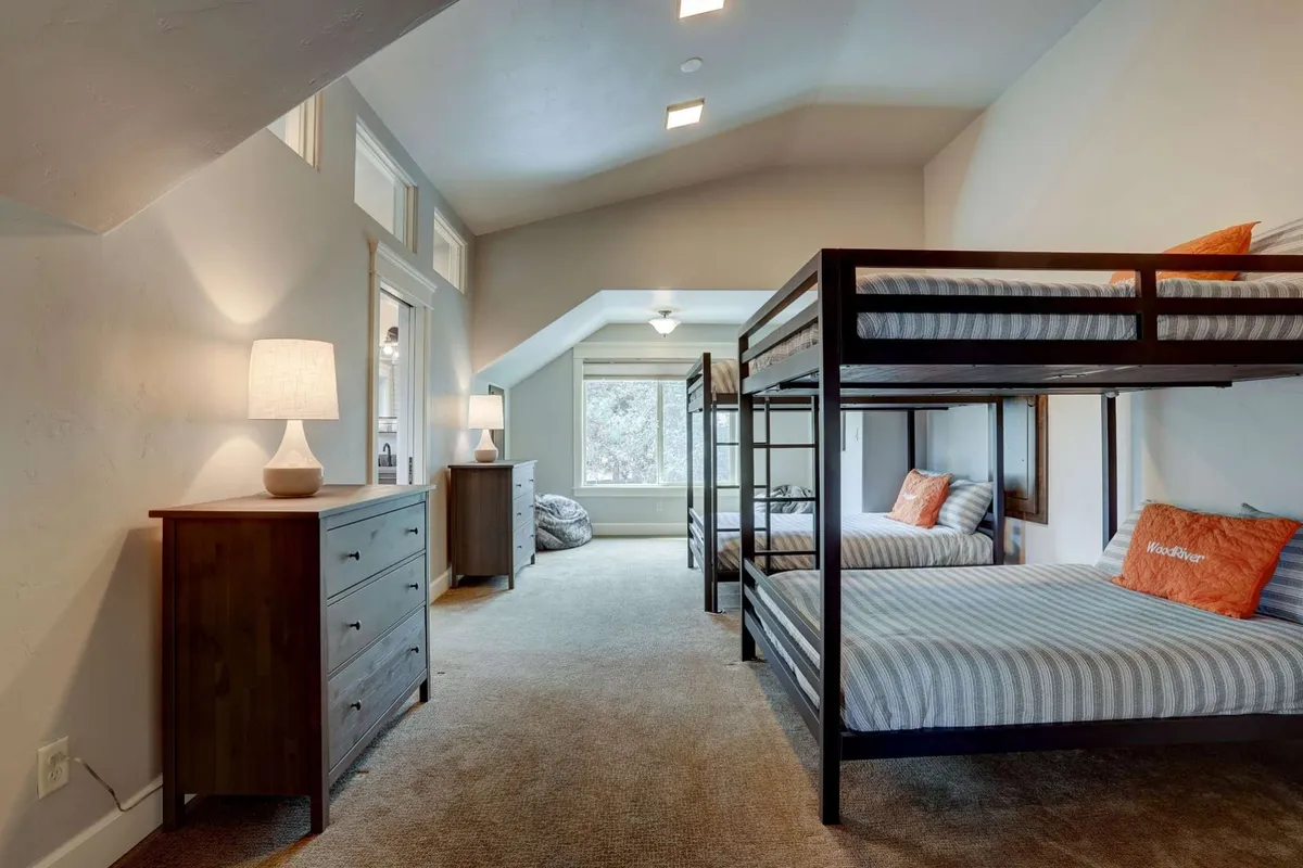 A playful and inviting bunk room featuring several bunk beds, a dresser, lamps, and a window that lets in plenty of light. The space feels cozy and ideal for kids or teens, with room for group activities and bonding.