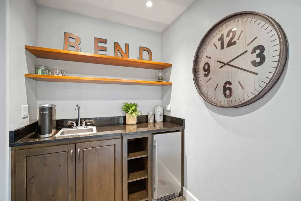 A compact kitchenette area highlighted by a distinct 'BEND' sign, a large wall clock, a sink, and a mini-fridge. The vibe is cozy and inviting, with touches that reference the local area and provide a setting for quick refreshments or morning coffee before heading out.