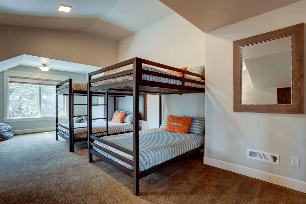 A bunk room filled with natural light, featuring bunk beds, accent pillows, a large window, and a decorative mirror. The space feels playful and is well-suited for group activities or sleepovers.
