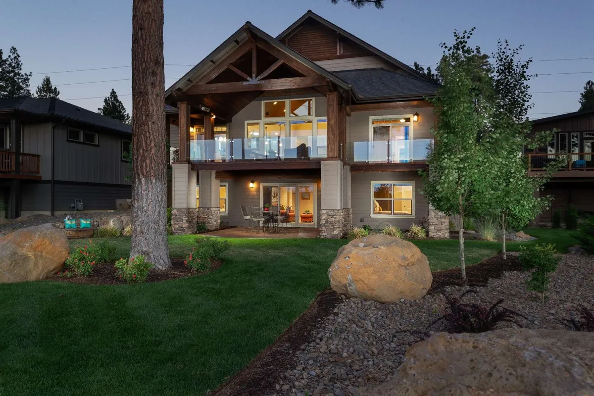 This image showcases the WoodRiver House's large front porch, illuminated windows, natural stone and wood accents, and a lush green lawn. The porch is inviting and hints at a welcoming transition between home and the outdoors, while the evening lighting evokes the idea of returning home after an adventure.