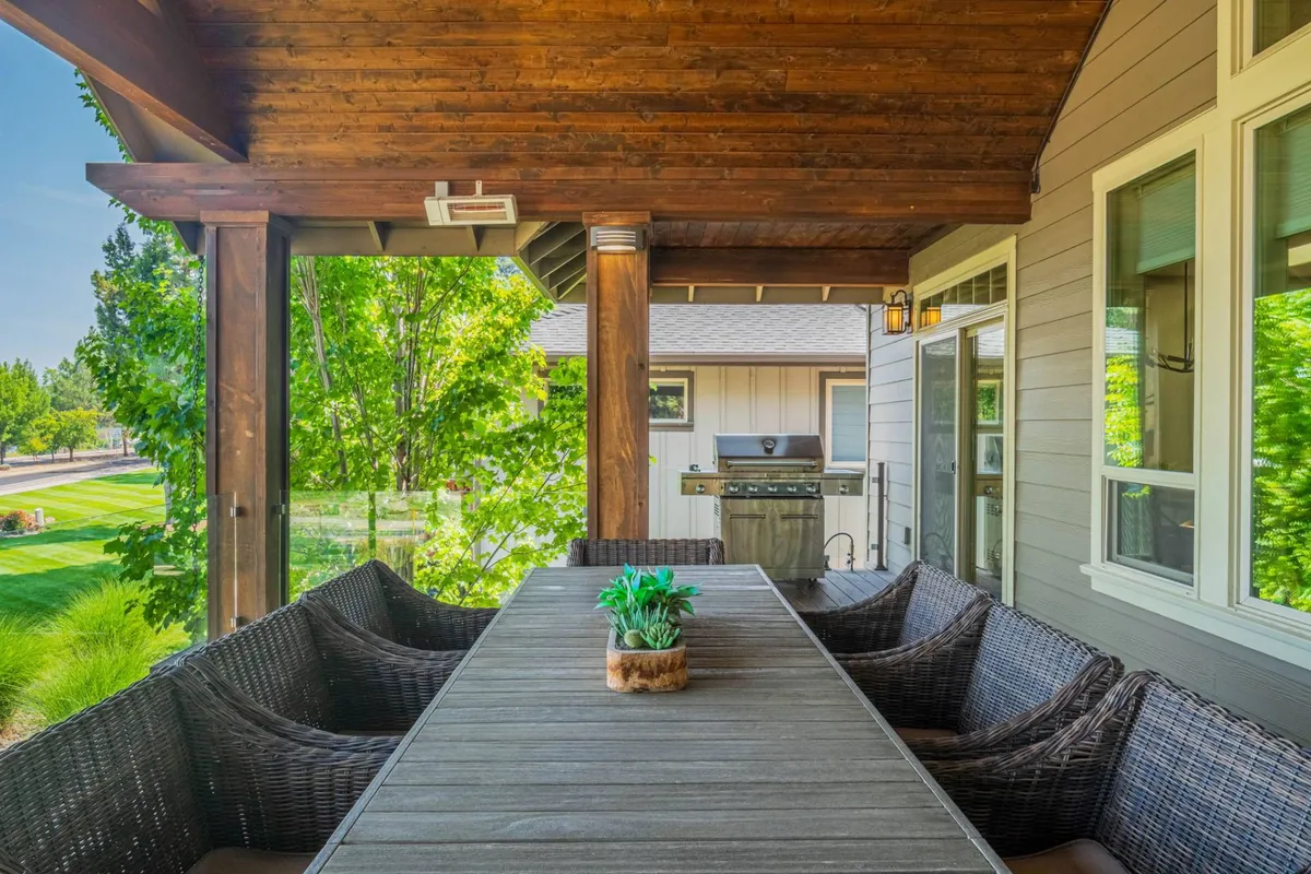 A long wooden dining table with woven chairs is set on a covered patio. Green plants and a grill are visible in the background, suggesting a space designed for outdoor dining and barbecues in all seasons.