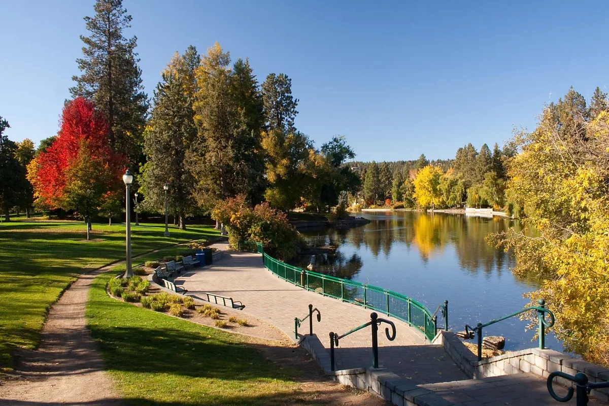 A serene view of the calm river bordered by vibrant trees and a winding walking path. A green railing indicates a lookout point, inviting residents to pause and take in the natural beauty of the Deschutes River.