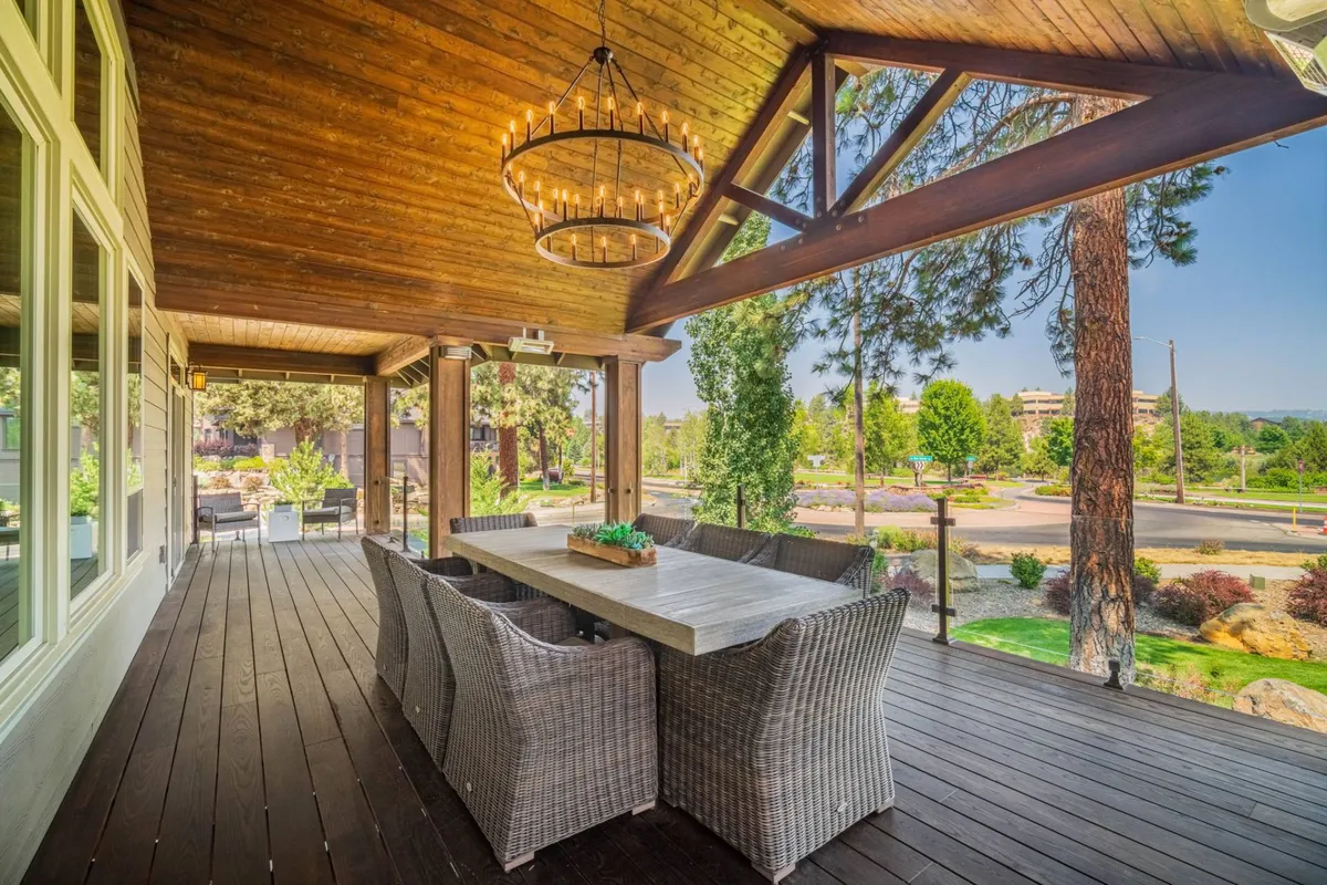 A spacious wooden deck is set for outdoor dining, featuring a large table beneath a glowing chandelier. The deck is surrounded by lush greenery, creating a tranquil atmosphere perfect for gatherings or relaxing in nature.